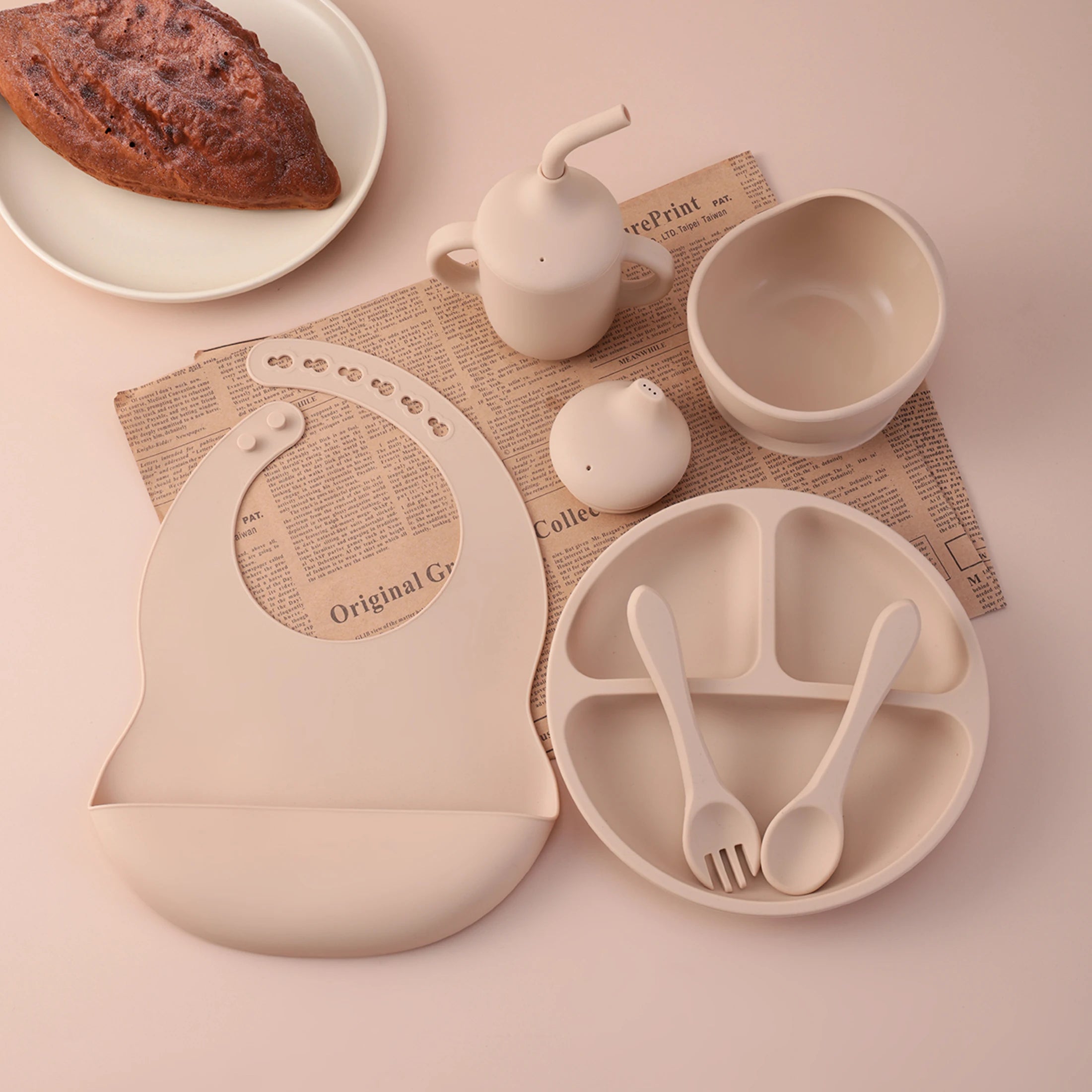 A coordinated baby feeding set in a warm cream tone arranged on a pale pink surface, including a divided plate with two utensils, a bib, a small teapot, a bowl, and a small container. A plate of bread is partially visible in the background as styling prop.