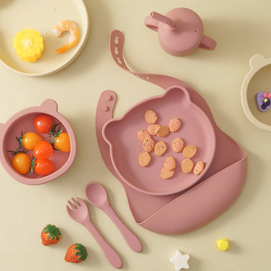 Pink children's tableware set including bowl, plate, and utensils on a beige background.