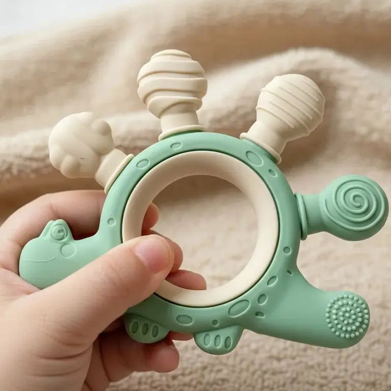 A baby teether toy held in a hand, featuring a mint green dinosaur design with cream-colored textured bumps and a ring handle, set against a soft beige blanket background.