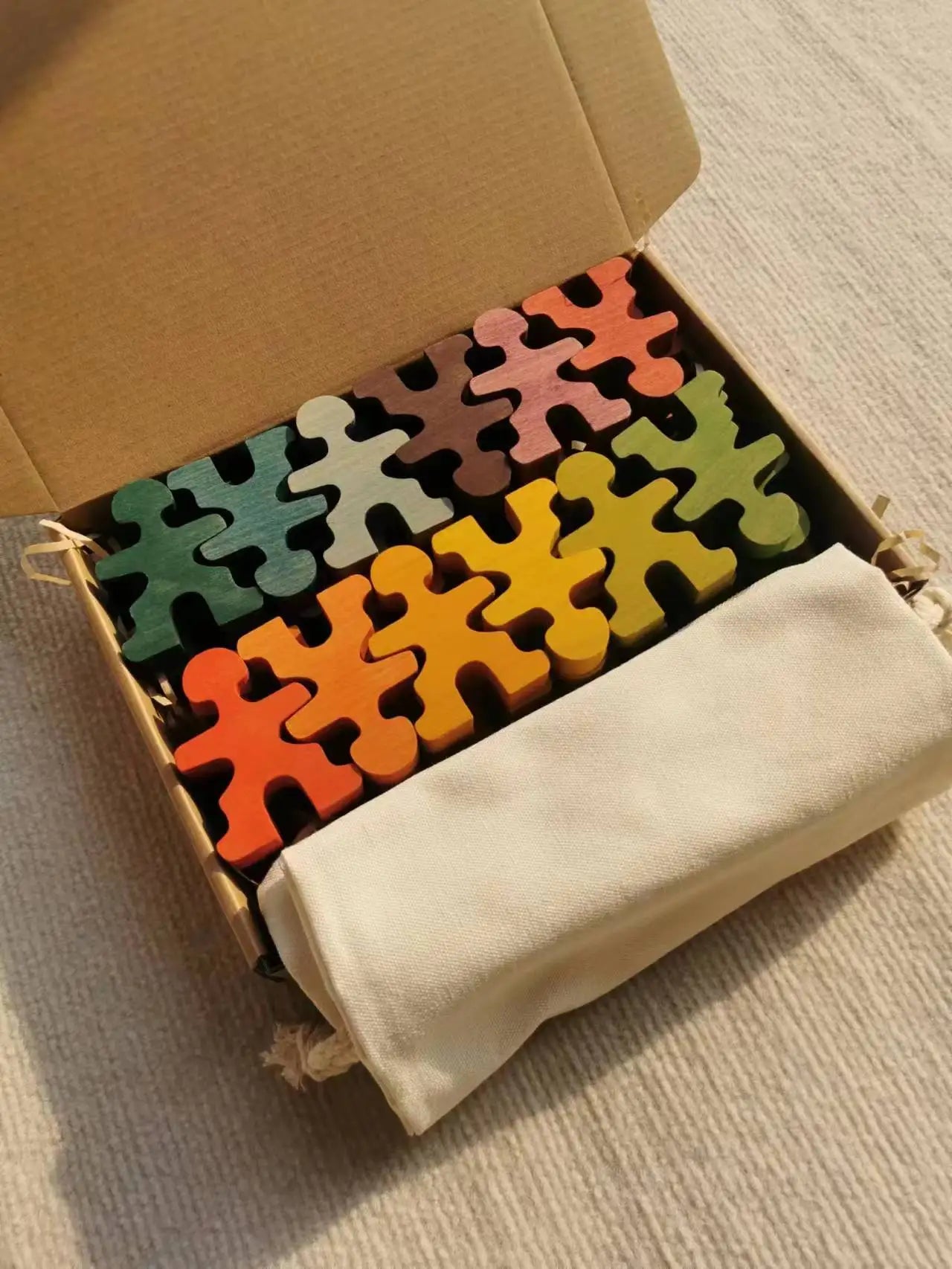 Colorful puzzle pieces in an open cardboard box with a beige drawstring bag on a light brown surface.