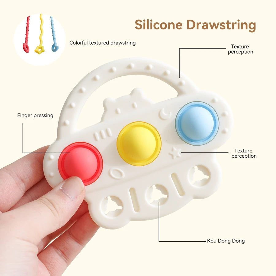 A hand holds a cream-colored silicone teething toy with three colorful textured buttons (red, yellow, and blue) and text callouts explaining features like finger pressing, texture perception, and kou dong dong, on a light beige background.