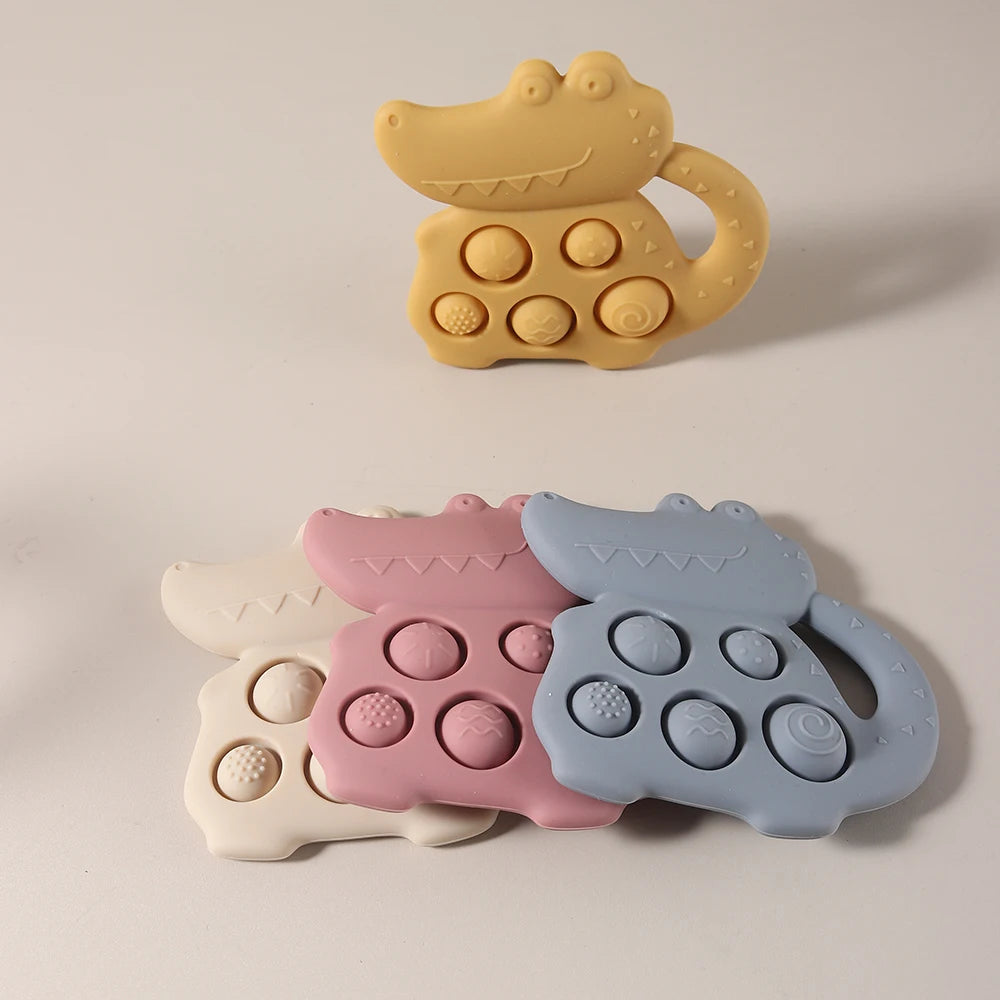 Set of four crocodile-shaped silicone teething toys in yellow, pink, and blue on a beige background.
