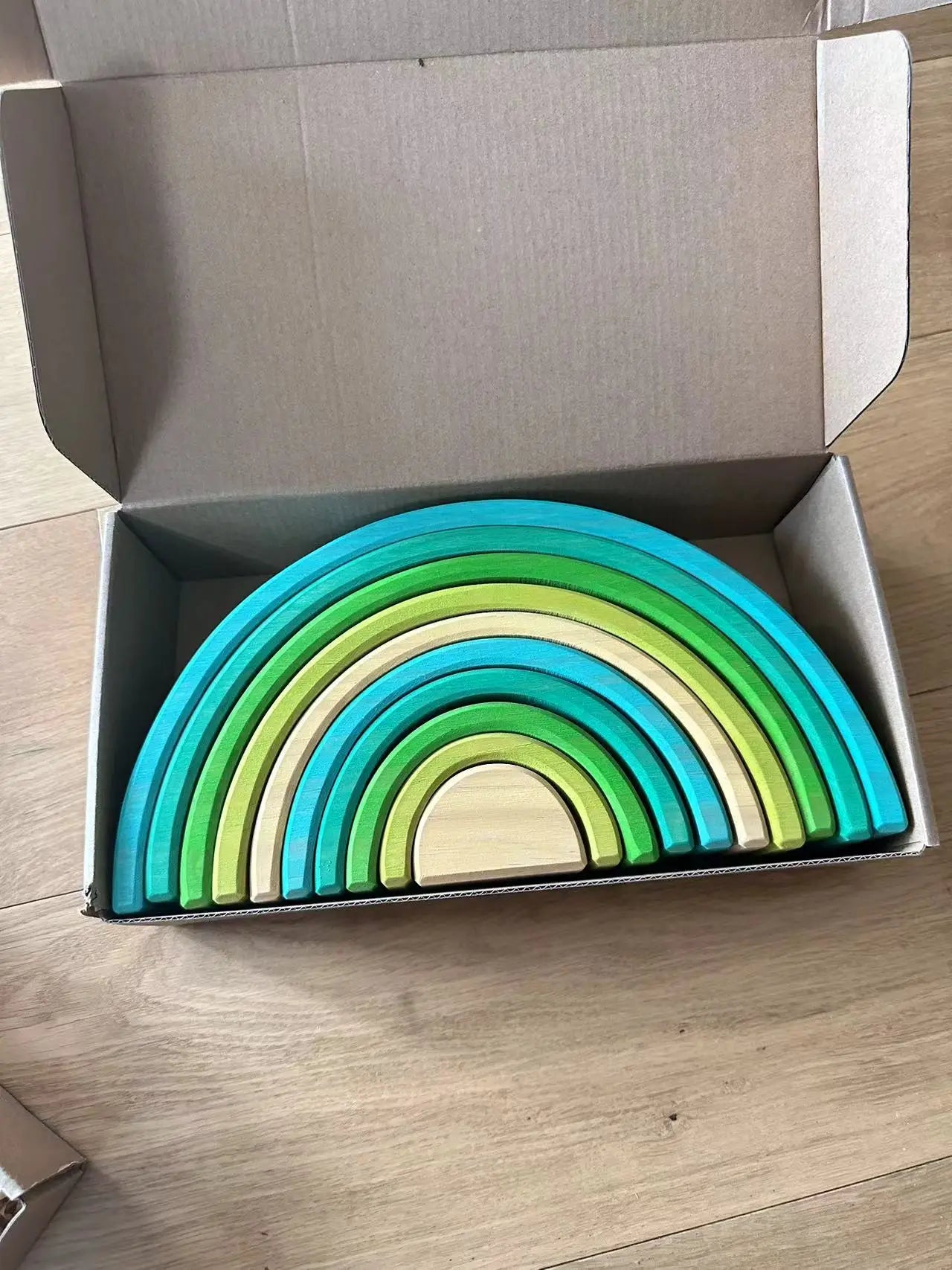 Set of colorful wooden rainbow toys in an open cardboard box on a wooden floor.