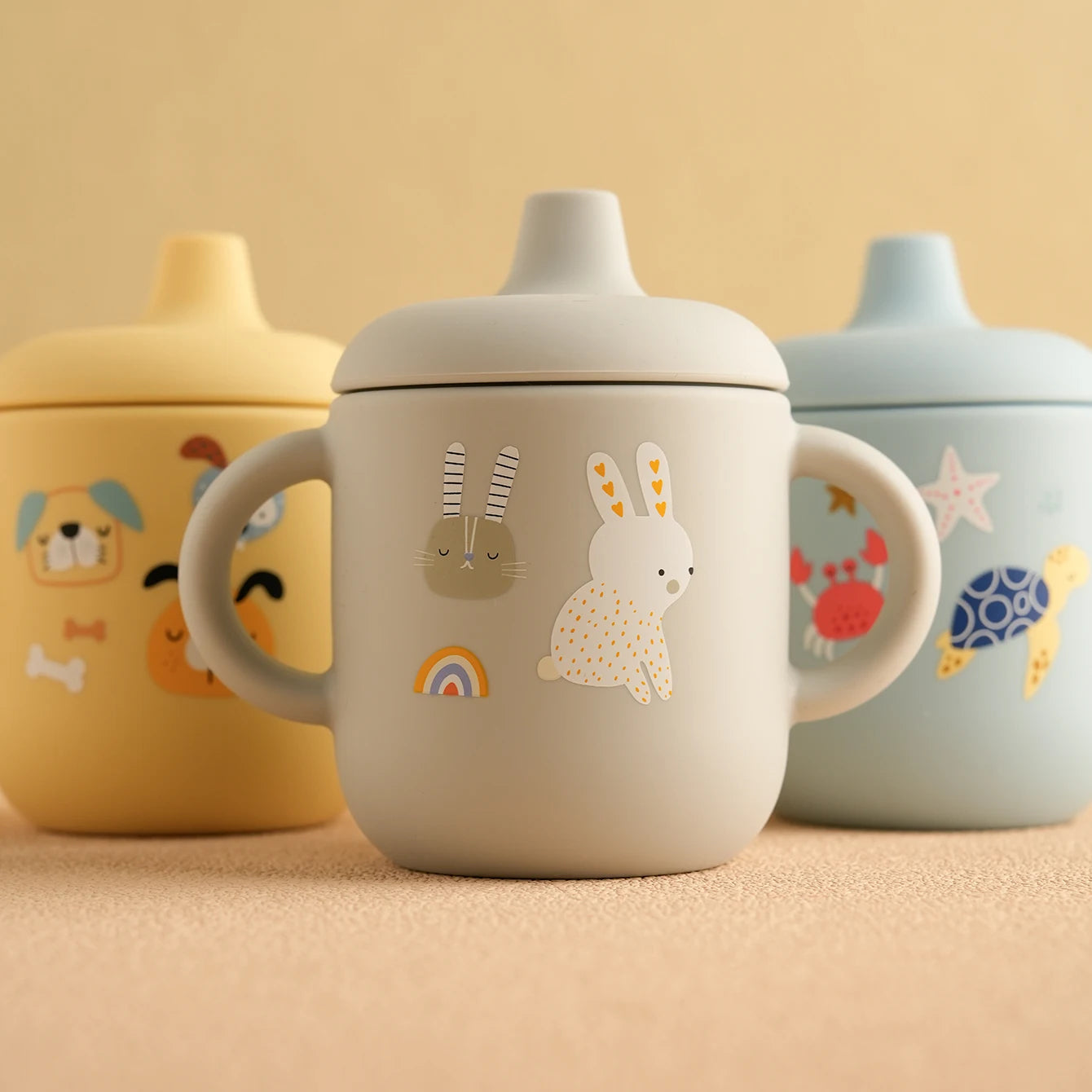 Cartoon print silicone sippy cups in yellow, grey and blue on a table