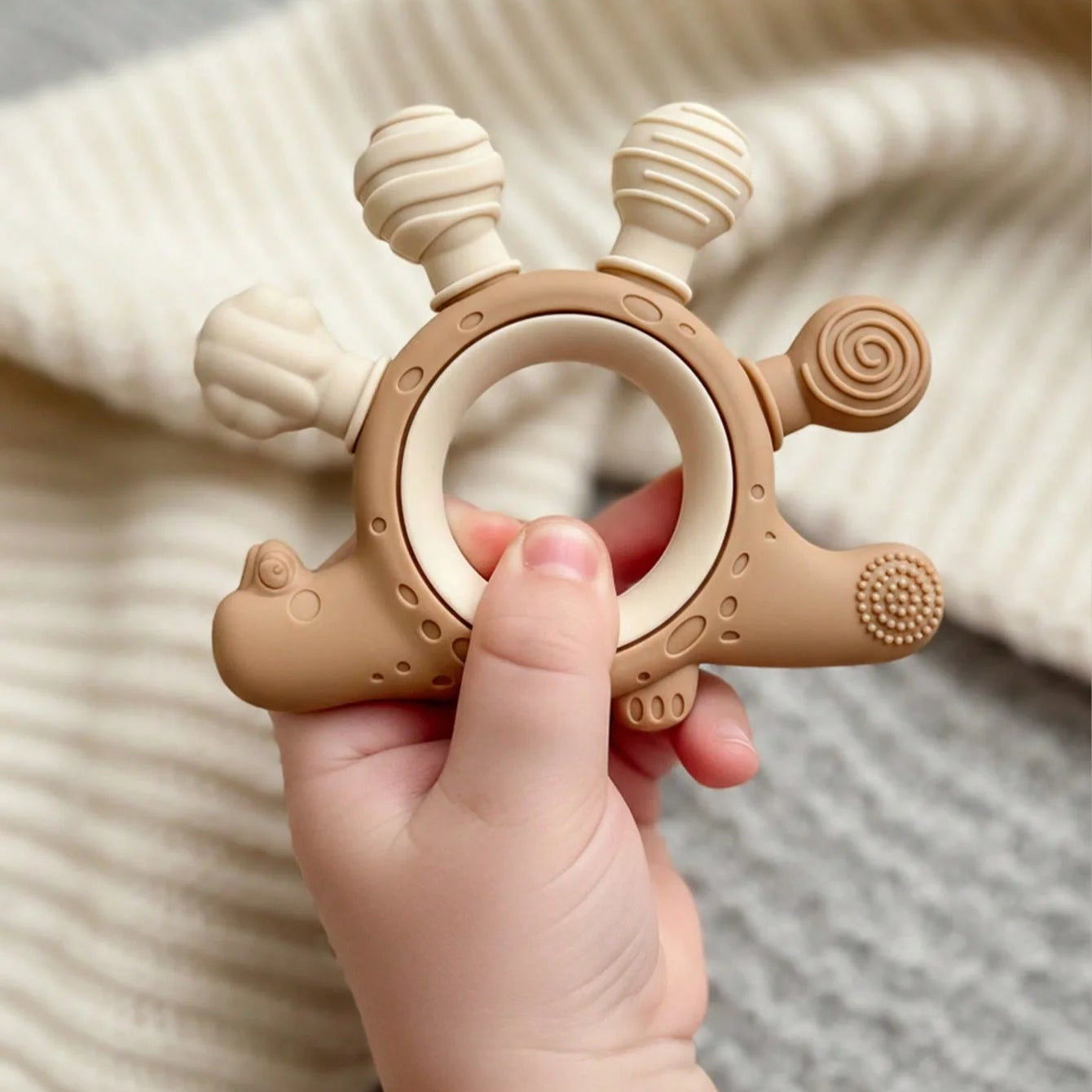 A baby teething ring toy held in a baby's hand, featuring a light brown silicone body with a wooden ring handle and textured sensory bumps, set against a soft cream knit blanket.