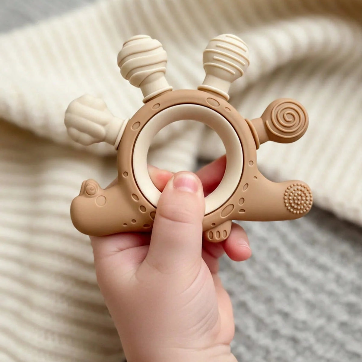 A baby teething ring toy held in a baby's hand, featuring a light brown silicone body with a wooden ring handle and textured sensory bumps, set against a soft cream knit blanket.