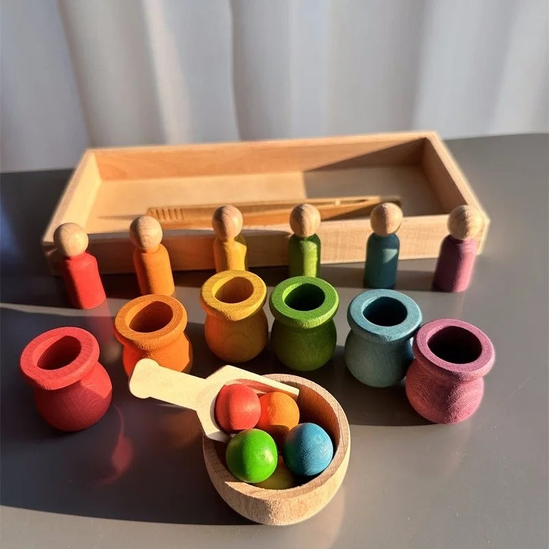 A colorful wooden Montessori-style sorting toy set arranged on a tabletop, including small wooden peg dolls, cylindrical color cups, a scoop, and a wooden tray, photographed in warm natural light.