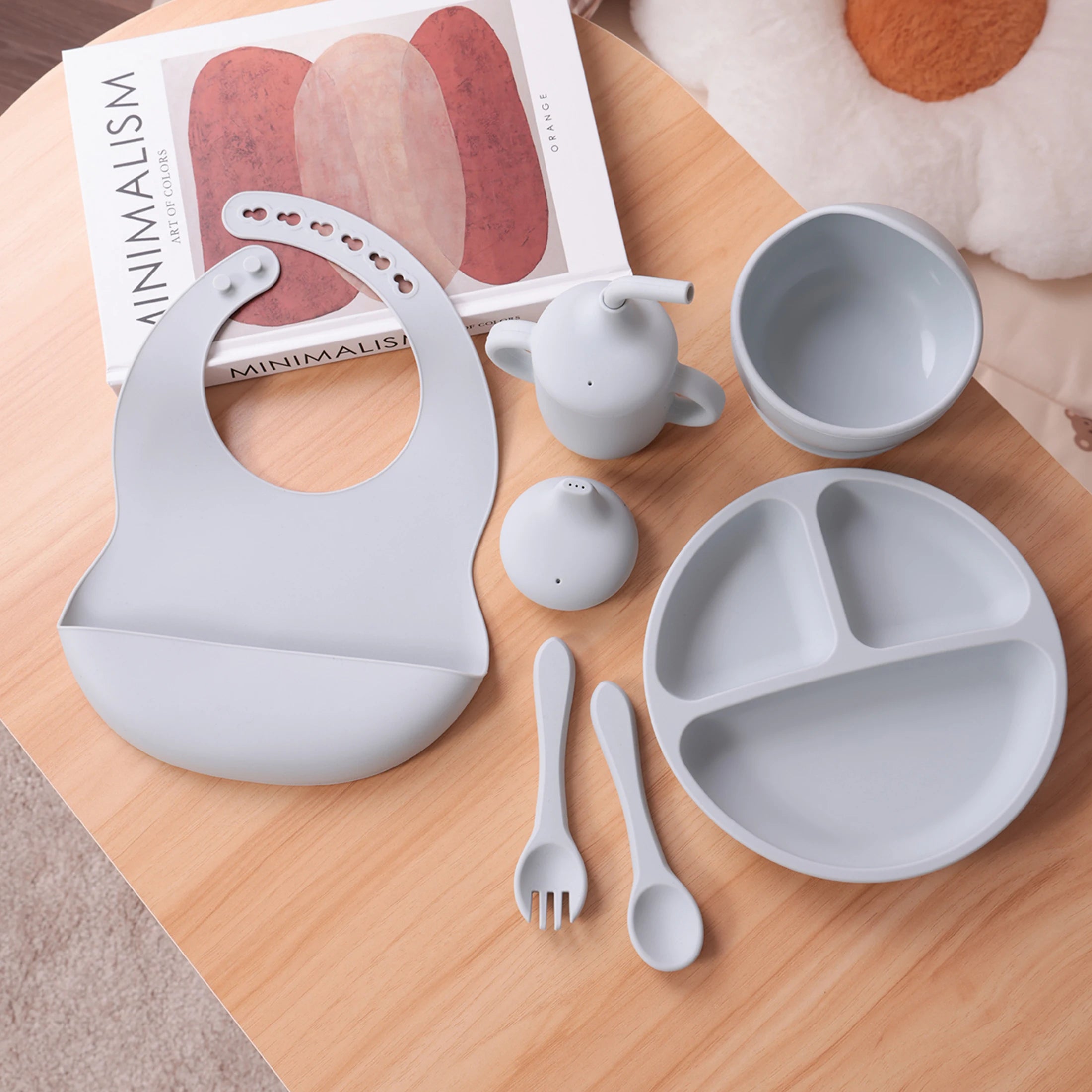 Set of baby feeding utensils including a bib, bowl, spoon, and fork on a wooden surface with a magazine in the background.