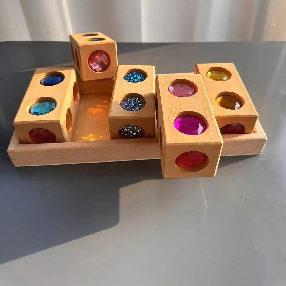 A wooden Montessori-style sorting toy with colorful gem-like circles in different colors, arranged on a tray on a gray surface with a soft curtain background.