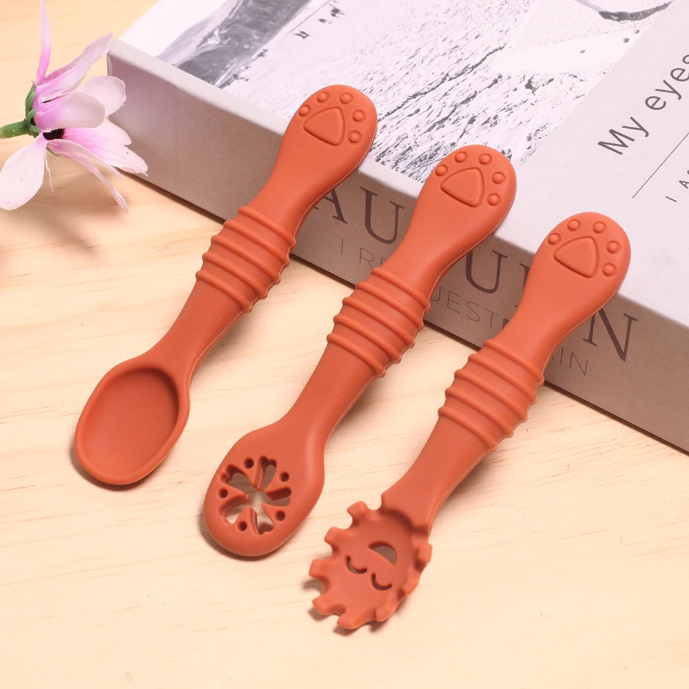 Three orange silicone baby spoons with unique designs on a light wooden surface.