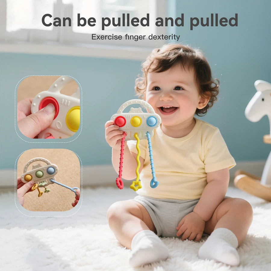 A baby sitting on a white rug holds a colorful baby toy with hanging pull strings. Two inset circles show close-ups of the toy being pulled by a hand.