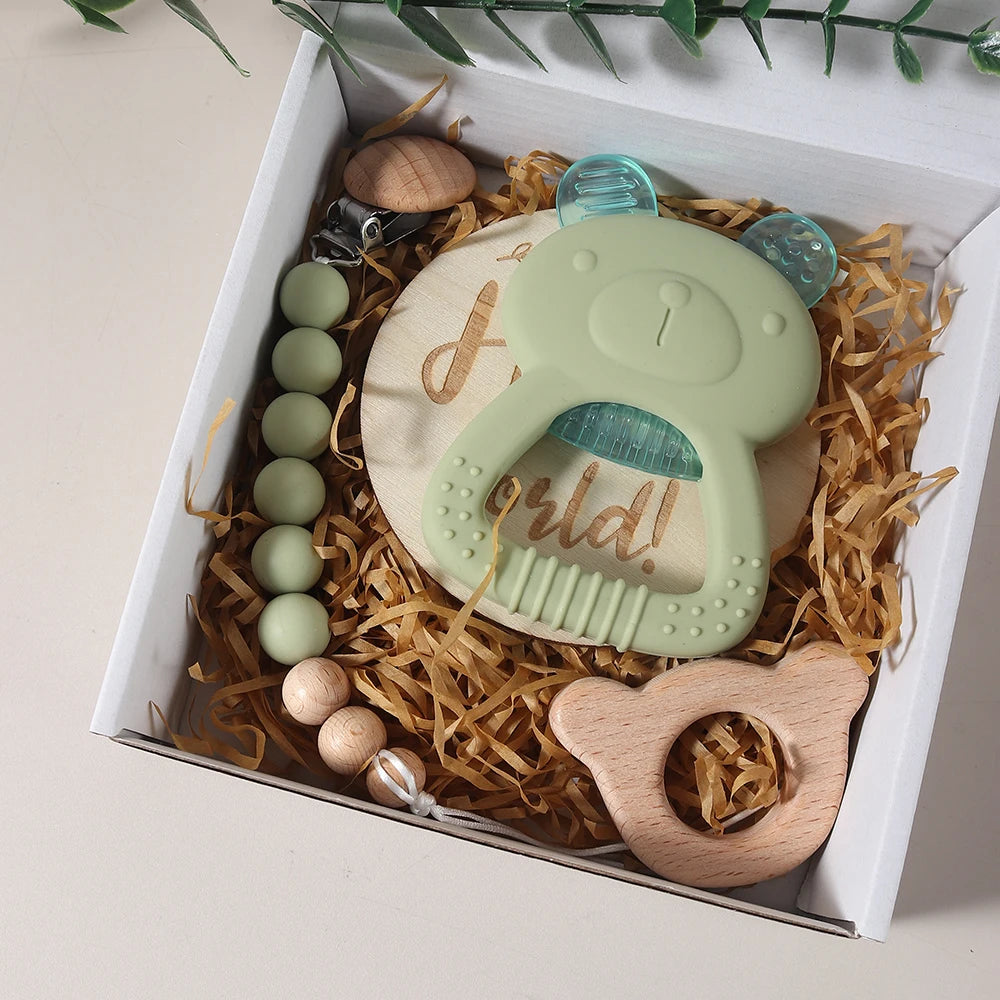 Set of baby toys including a green teether, wooden beads, and a wooden rattle in a box with straw.