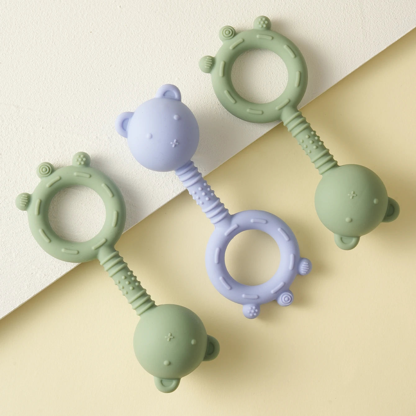 Two silicone teething rings, one green and one purple, on a beige background.