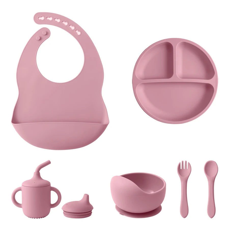 A pink silicone baby feeding set on a white background, including a bib, divided plate, sippy cup with lid, bowl with suction base, and utensils (fork and spoon).