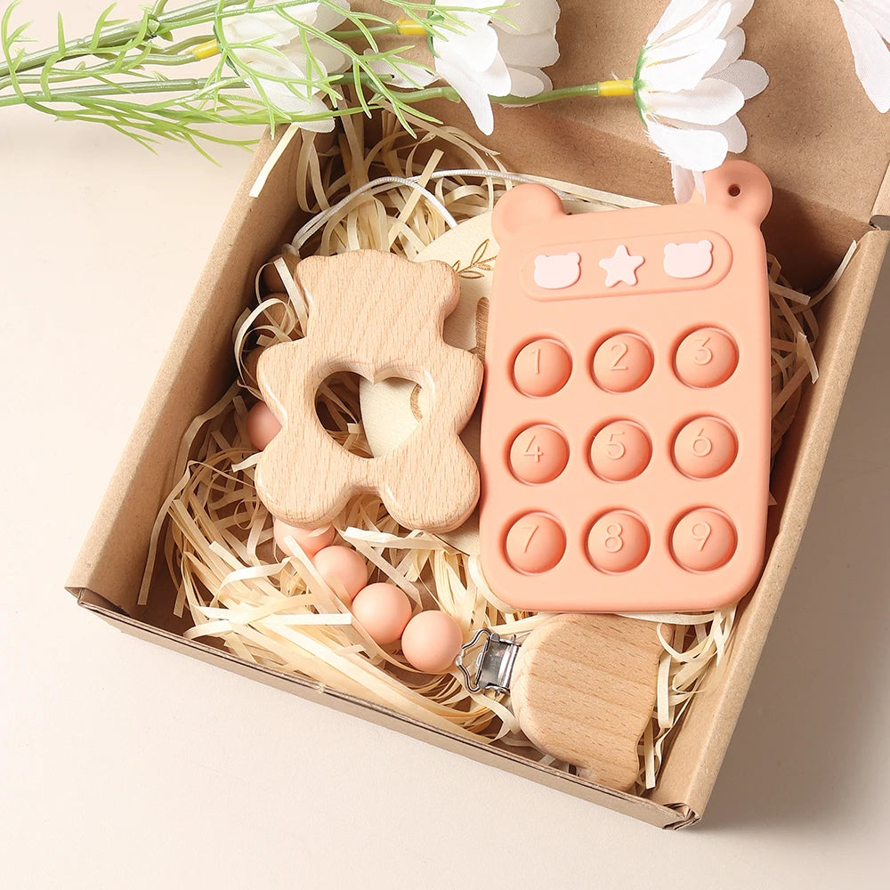Baby teething toys including a wooden bear, pink phone, and wooden brush in a decorative box with flowers.