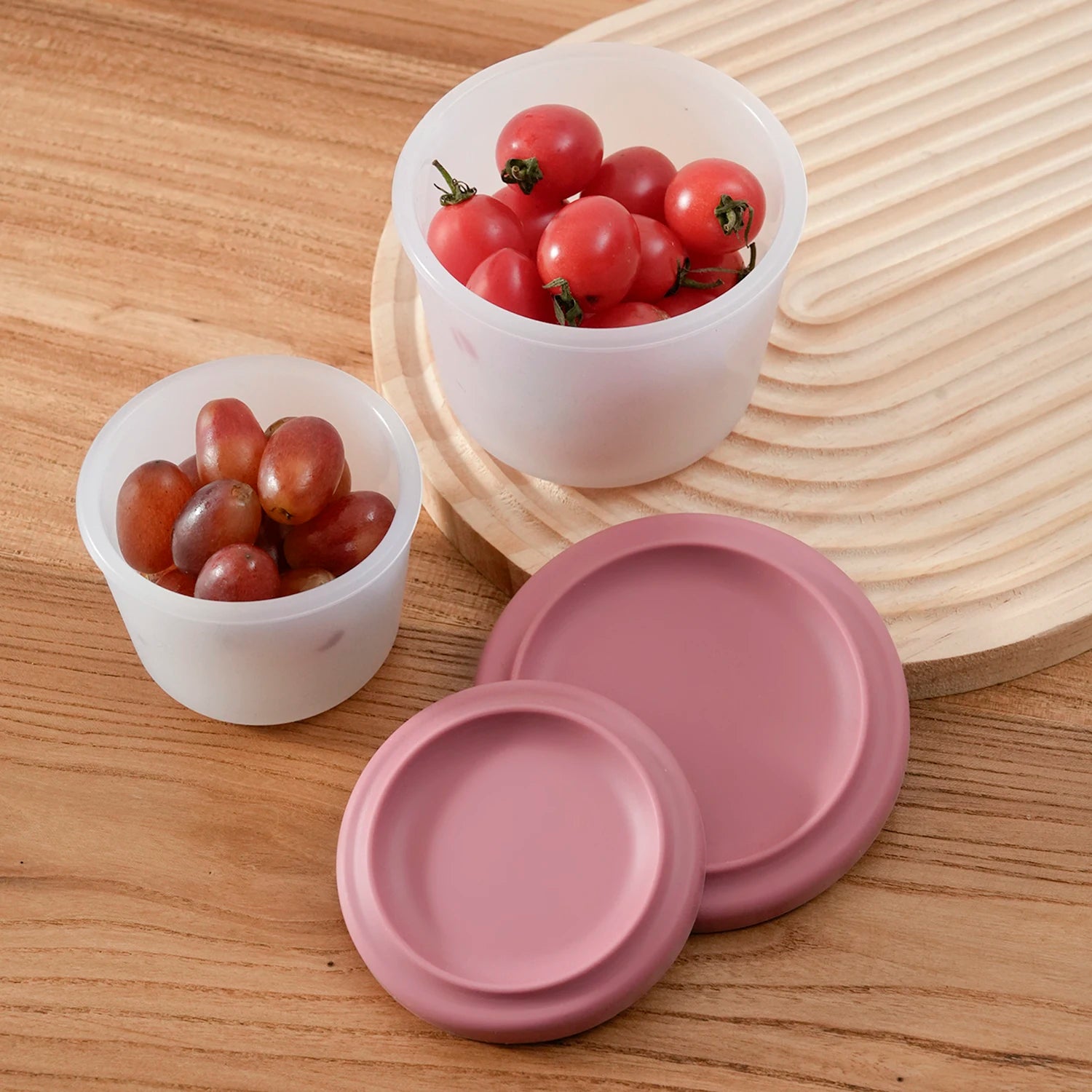Two round silicone containers with tomatoes and grapes beside their rose lids