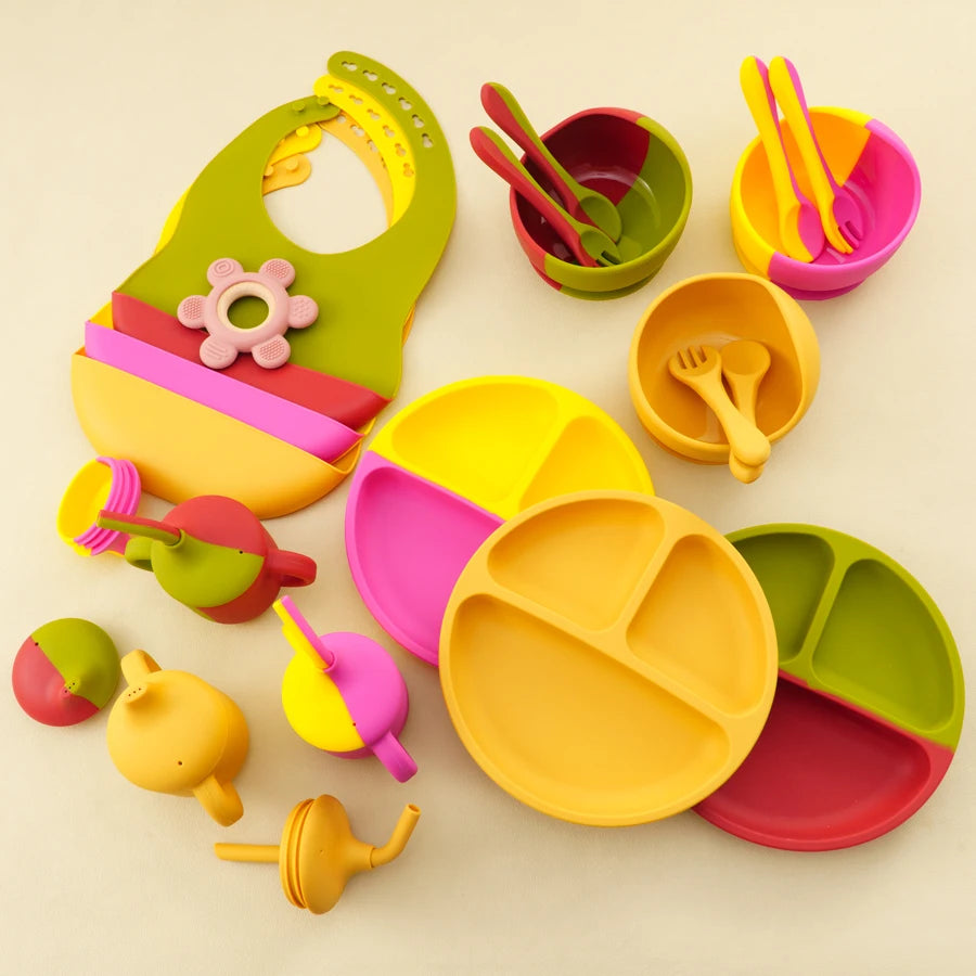 A colorful children's play mealtime set arranged on a light beige background, including divided plates, cups, spoons, utensils, and a bib with a flower detail.