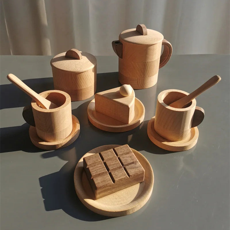 Set of wooden kitchen utensils including a teapot, cups, and a knife on a gray surface with a white curtain background.