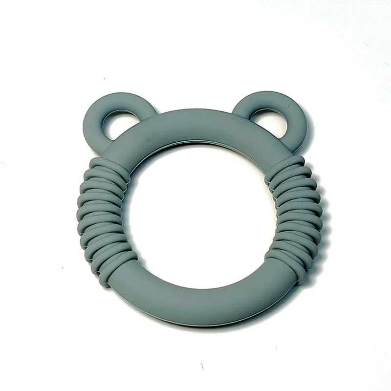 A gray silicone baby teether with a circular design, featuring a textured, ribbed outer band and two small loop handles resembling ears at the top, shown on a plain white background.
