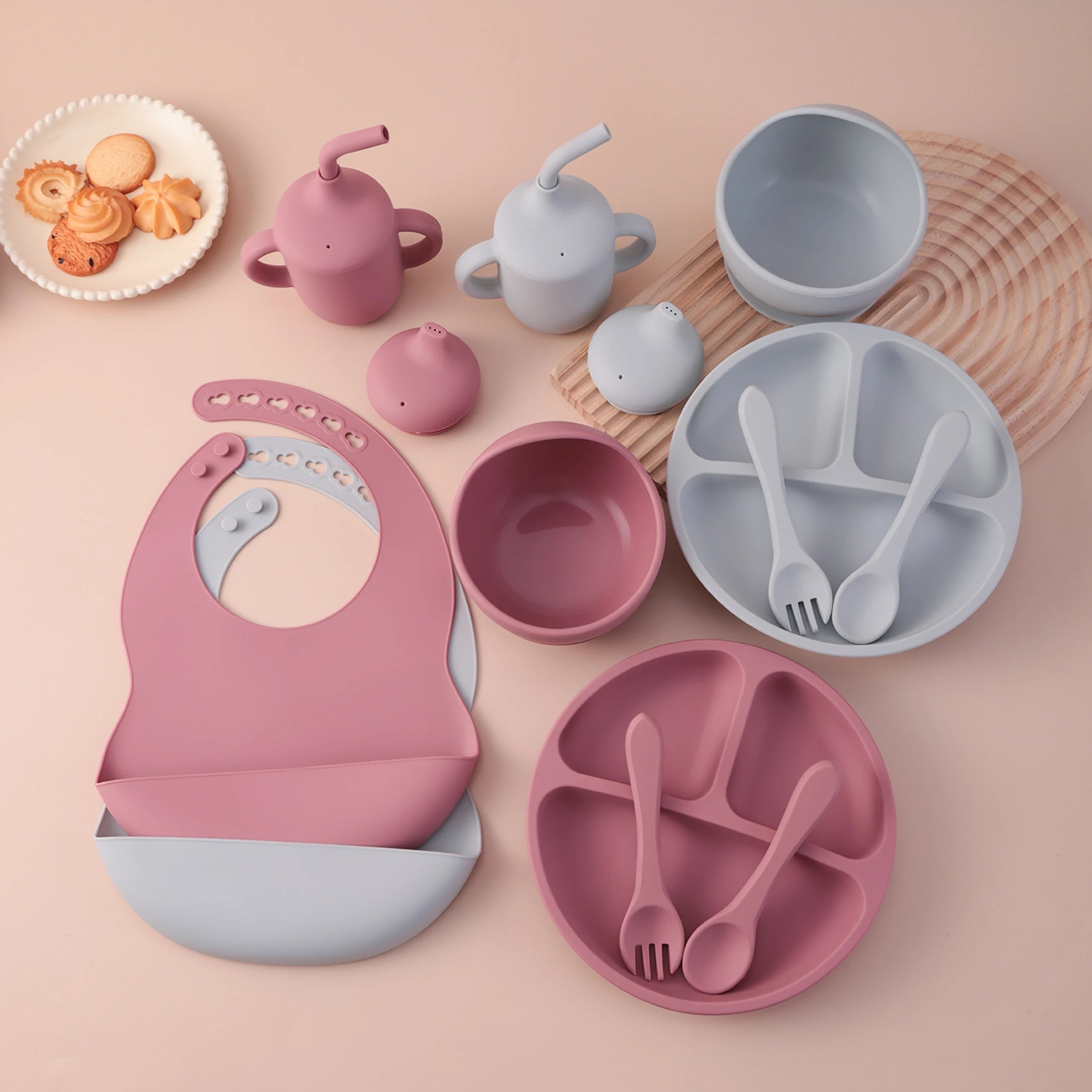 A coordinated baby feeding set arranged on a soft blush background, including a pink bib, two sippy cups, a round divided plate, a bowl, small bowls, and utensils in matching pink and pale blue-gray tones. A small plate of cookies appears at the upper left as a prop.