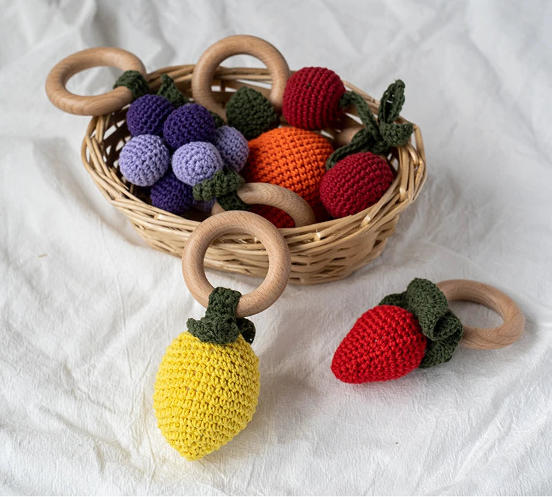 Crocheted fruit toys with wooden rings on a white fabric background, branded 'OURSHOURS'.