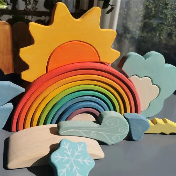 A set of colorful wooden toy blocks shaped like a rainbow, sun, clouds, mountains, and a snowflake, arranged on a tabletop in natural sunlight.