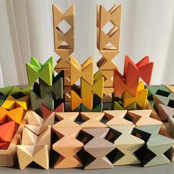 A collection of colorful wooden toy blocks arranged in a pyramid-like display. The blocks are made of natural wood with painted accents in green, yellow, orange, red, and gray, featuring geometric and star-like shapes.